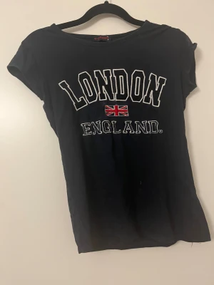 Svart London England t-shirt G-Crown - Svart t-shirt från G-Crown med tryck 'LONDON ENGLAND' och brittiska flaggan på bröstet. Klassisk rund hals och korta ärmar. Skön och enkel modell i bomull, perfekt för dig som gillar streetstyle och cityvibe.