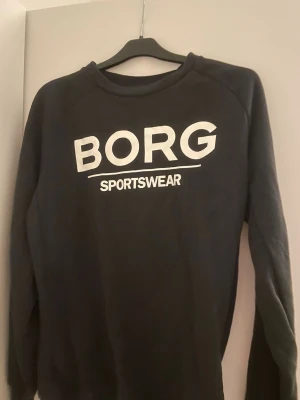 Svart sweatshirt från Björn Borg - Svart sweatshirt från Björn Borg med stor vit logga 'BORG SPORTSWEAR' på bröstet. Tröjan har rund hals och långa ärmar, tillverkad i mjuk bomull som är perfekt för chill eller träning. Klassisk och enkel design som funkar till jeans eller joggers.