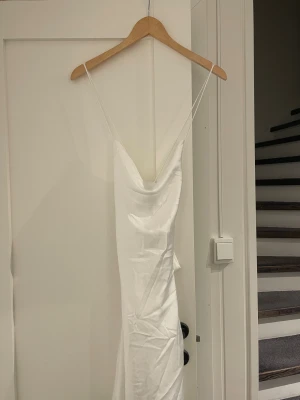 Vit långklänning med smala axelband - Säljer en elegant vit långklänning med smala axelband som korsas i ryggen. Klänningen har en snygg draperad halsringning och är tillverkad i ett glansigt satinliknande material. Perfekt för dig som vill ha en stilren och chic look.
