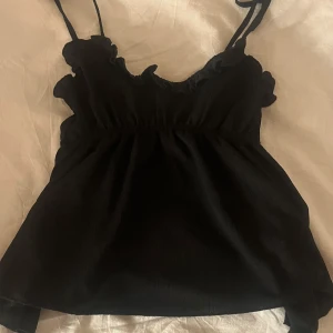 Svart linnetopp med volangkant - Säljer en svart linnetopp med smala axelband och volangdetaljer längs halsringningen. Toppen har en lätt rynkad midja som ger en söt och feminin siluett. Perfekt för dig som gillar enkel men ändå stylish design.
