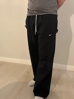 Nike Solo Swoosh storlek L - Svarta mjukisbyxor från Nike med vit snörning i midjan och diskret Nike-logga på vänster ben. Byxorna har en loose passform och raka ben, tillverkade i mjukt bomullsmaterial. Perfekt för chill eller träning, storlek L.