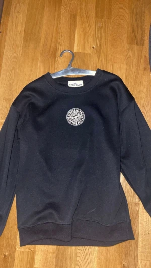 Svart Stone Island sweatshirt - Svart sweatshirt från Stone Island med vit logga tryckt på bröstet och stort tryck på ryggen. Tröjan är andvänd typ 3 gånger den är nästan i helt nytt skick.