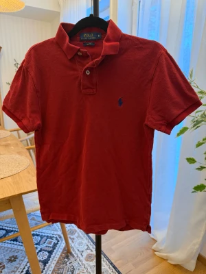 Röd Polo Ralph Lauren piké slim fit - Snygg röd pikétröja från Polo Ralph Lauren i slim fit-modell. Klassisk krage, korta ärmar och två knappar framtill. Ikoniska blå loggan broderad på bröstet. Perfekt för dig som gillar stilrena och sportiga plagg.