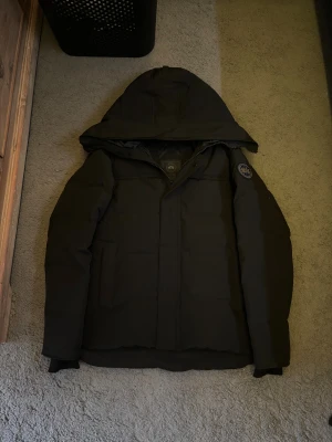 Canada Goose jacka - Säljer en svart pufferjacka från Canada Goose i storlek S. Jackan har huva, diskret logotyp-patch på ärmen och dragkedja framtill. Fodrad för extra värme och perfekt för kalla dagar. Klassisk, stilren design med raka linjer och två sidofickor.