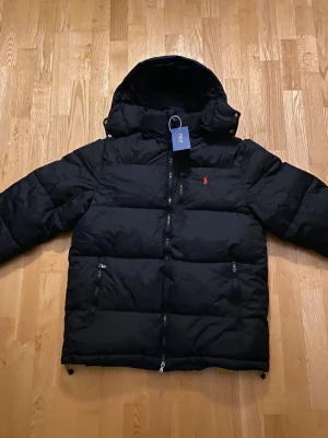 Svart pufferjacka från Polo Ralph Lauren - Säljer en svart pufferjacka från Polo Ralph Lauren med huva och dragkedja. Jackan har det klassiska röda Polo-logot på bröstet och två sidofickor med dragkedja. Perfekt för kalla dagar och riktigt snygg till streetwear-stilen. Levereras packad i påsen som det syns i bilden. Storlek M. 