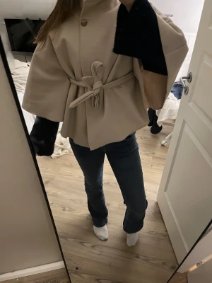 Beige poncho Missäy  - Säljer en beige kappa/poncho med knytbälte i midjan och vida trekvartsärmar. Jackan har en knapp upptill och en snygg, oversized passform som ger en chill vibe. Perfekt för dig som vill ha en stilren och trendig look.