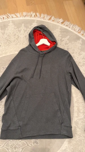 Grå hoodie från Boss med röd huva - Snygg grå hoodie från Boss med diskret logga på bröstet. Insidan av huvan är röd vilket ger en cool kontrast. Tröjan har snörning vid huvan, känguruficka och långa ärmar. Perfekt för chill dagar och streetstyle.