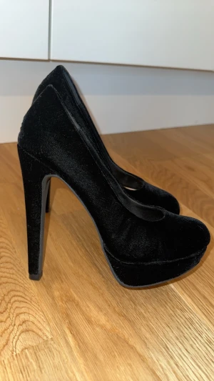 Svarta sammetsklackar från H&M - Snygga svarta pumps från H&M i storlek 36, tillverkade i mjuk sammet med hög klack och plattform. Skorna har rund tå och en stilren, elegant look som passar perfekt när du vill sticka ut lite extra. Perfekt för dig som gillar klassisk stil med en twist.
