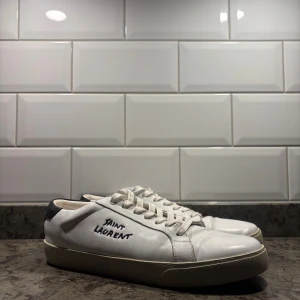 Vita Saint Laurent skor - Snygga vita sneakers från Saint Laurent med klassiska logga på sidan. Skorna har rund tå, platt sula och snörning framtill. Hälpartiet är svart och resten av skon är vit, tillverkad i mjukt skinn. Mycket snygga till t.ex kavaj eller kostym. Pris går att förhandlas vid snabb affär👍