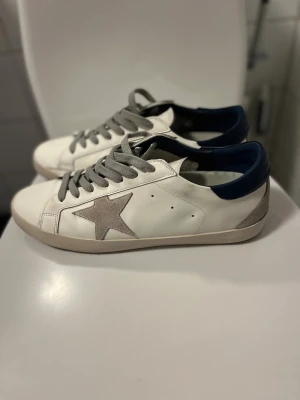 Golden Goose Superstar sneakers - Golden Goose Superstar sneakers i vitt skinn med grå mockastjärna på sidan, grå snörning och mörkblå häl. Klassisk rund tå och platt sula. Snygg kontrast mellan färgerna och den ikoniska stjärndetaljen gör dem riktigt trendiga.