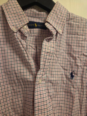 Rutig skjorta från Ralph Lauren - Säljer en klassisk rutig skjorta från Ralph Lauren i bomull. Skjortan har lång ärm, knappar framtill och en broderad marinblå logga på bröstet. Färgerna är ljusrosa, vit och blå i ett rutigt mönster. Perfekt för dig som gillar stilrena och tidlösa plagg.