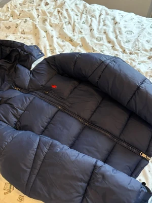 Mörkblå pufferjacka från Polo Ralph Lauren - Säljer en mörkblå pufferjacka från Polo Ralph Lauren med röd logga på bröstet. Jackan har dragkedja framtill och två sidofickor med dragkedja. Materialet är polyester och jackan är quiltad för extra värme. Perfekt för kalla dagar!