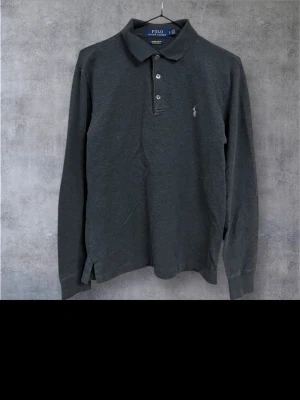 Mörkgrå långärmad polotopp från Polo Ralph Lauren - Säljer en mörkgrå långärmad polotopp från Polo Ralph Lauren med klassisk krage och tre knappar framtill. Toppen har en broderad logga på bröstet och är tillverkad i mjuk bomull. Perfekt för en stilren och avslappnad look.