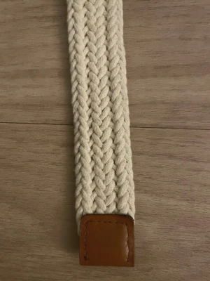Beige flätad bälte med skinn-detaljer - Snyggt flätat bälte i beige med bruna skinn-detaljer vid spännet och ändarna. Bältet har ett klassiskt metallspänne och är gjort i ett grovt bomullsmaterial som ger en cool och avslappnad vibe. Perfekt för att lyfta din outfit med en enkel men stilren accessoar.