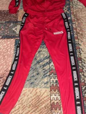 Röda mjukisbyxor från Hoodrich - Snygga röda mjukisbyxor från Hoodrich med svarta sidoremsor där det står 'HOODRICH' i vitt. Byxorna har snörning i midjan och en slim passform som sitter tajt längs benen. Perfekta för chill eller streetwear-stil.