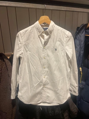 Vit skjorta från Polo Ralph Lauren - Vit skjorta från Polo Ralph Lauren i storlek 16 med klassisk krage och knappar framtill. Broderad marinblå logga på bröstet. Långärmad och tillverkad i mjuk bomull. Slim passform som ger en clean och stilren look. Perfekt för dig som gillar tidlös design och snygga detaljer. ny pris: 1100kr
