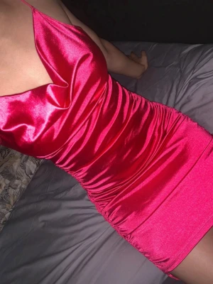 Rosa satin kortklänning med drapering - Säljer en glansig rosa kortklänning i satin med smala axelband och snygg draperad urringning. Klänningen har en tight passform och är perfekt för dig som vill sticka ut med en färgstark look. Supersnygg att styla med klackar eller boots.