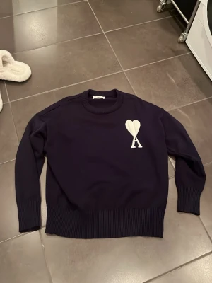 Mörkblå stickad tröja från Ami - Snygg mörkblå stickad tröja från Ami med vit hjärtapplikation och bokstaven A på bröstet. Tröjan har rund hals, ribbade muddar och är långärmad. Perfekt för dig som gillar stilrena detaljer och vill ha något som sticker ut lite.