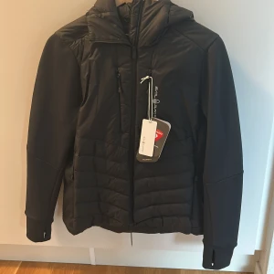 Svart pufferjacka Sail Racing M - Svart pufferjacka från Sail Racing i storlek medium. Jackan har quiltad framsida och rygg, huva med logga och dragkedja framtill. Ärmarna är släta och har tumhål vid mudden. Perfekt för kyliga dagar och snygg till streetwear.