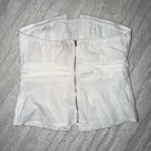 Vit korsett-topp med dragkedja - Säljer en vit korsett-topp med dragkedja framtill. Toppen är axelbandslös och har en stilren, strukturerad form med elastisk kant upptill för bättre passform. Perfekt att styla med jeans eller kjol för en trendig look.