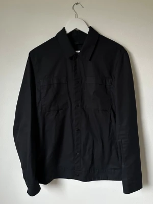 Svart overshirt från Selected Homme - Svart overshirt från Selected Homme i ekologisk bomull. Jackan har klassisk krage, två stora bröstfickor med knappar och långa ärmar med manschett. Perfekt att slänga på över en hoodie eller t-shirt för en clean look. 5M2