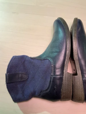 Mörkblå boots från Clarks - Säljer ett par mörkblå boots från Clarks med snyggt mönstrad textil upptill och slät skinnfinish nedtill. Skorna har låg klack, rund tå och robust sula. Perfekta för dig som gillar en mix av material och stilrena detaljer.läder, äkta.