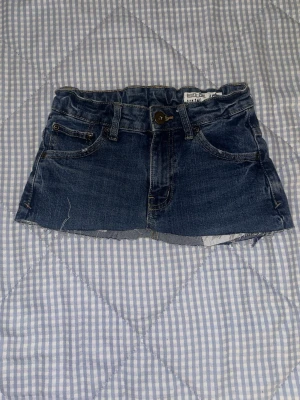 Mörkblå jeanskjol med rå kant - Säljer en mörkblå jeanskjol från Shorty med låg midja och rå, fransig kant nedtill. Kjolen har klassiska jeansdetaljer som fickor fram och bak, knapp och dragkedja. Perfekt för dig som gillar en edgy och avslappnad stil.mini skirt från 2000s