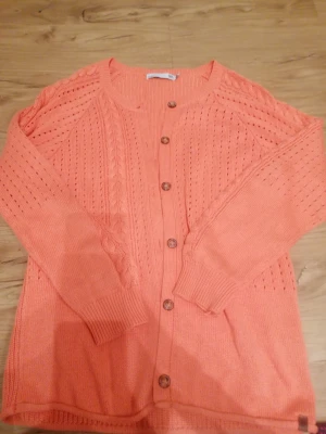 Korallfärgad stickad kofta från Lager 157  - Snygg korallfärgad stickad kofta från LGT med dekorativa knappar framtill. Koftan har långärmade ärmar, ribbade muddar och ett snyggt hålmönster samt flätstickade detaljer. Perfekt för lager-på-lager under kyliga dagar. Den har två fläckar, en vid armen & en vid bröstet. 😊