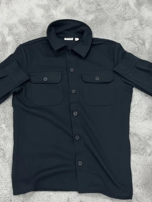Svart overshirt från Only & Sons - Snygg marinblå overshirt från Only & Sons med klassisk krage och två stora bröstfickor med knappar. Skjortan har långa ärmar och knäppning hela vägen framtill. Perfekt att slänga på över en t-shirt för en chill look. Pris kan diskuteras då den är oanvänd