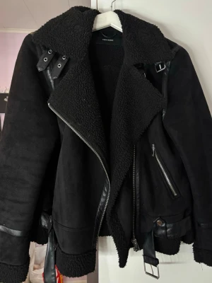 Svart teddyjacka från Vero Moda - Säljer en svart teddyjacka från Vero Moda med bred krage, detaljer i skinnimitation och grova dragkedjor. Jackan har bälte vid midjan och  spännen vid kragen. 