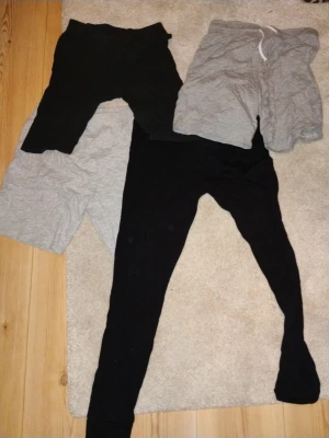 Tre mjukisshorts och leggings - Säljer ett paket med tre byxor: två mjukisbyxor i grått och svart, samt ett par svarta leggings. De grå mjukisbyxorna har snörning i midjan och är kortare, medan de svarta leggingsen är långa och tighta. Perfekt för chill eller träning.