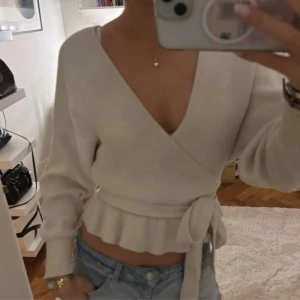 Ljusbeige omlottstickad tröja - Säljer en ljusbeige omlottstickad tröja med v-ringning och volangkant nedtill. Tröjan har lång ärm, knyt i midjan och öppen rygg för en snygg och trendig look. Perfekt att matcha med jeans eller kjol för en chill stil.