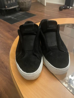 Svarta sneakers från Axel Arigato - Svarta sneakers från Axel Arigato i mocka med vit platt sula. Diskret guldtext på sidan och minimalistisk design utan snörning. Skorna har rund tå och är perfekta för en clean och stilren look. Säljer dem för att jag inte använder dem längre, priser kan diskuteras vid snabbaffär!!!!