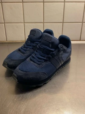 Mörkblå sneakers från Hugo Boss - Snygga mörkblå sneakers från Hugo Boss med mesh och mockadetaljer. Skorna har snörning, rund tå och en platt sula med tydlig Boss-logga undertill. Perfekt för dig som gillar stilrena och sportiga skor med coola materialmixar.