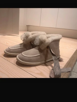 Beige UGG boots med pärldetaljer - Säljer ett par  UGG boots med fluffig päls runt kanten och breda band med UGG-logga DIREKR FRÅN DUBAJ. Skorna har pärldetaljer på snörningen och är tillverkade i mjuk mocka med platt sula. Perfekta för kalla dagar och har en rund tå.
