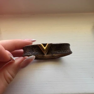 Brun Louis Vuitton läderarmband - Snyggt brunt armband från Louis Vuitton i äkta läder med klassiskt LV-mönster och guldfärgad V-detalj framtill. Armbandet har justerbar knäppning med två guldfärgade nitar och är perfekt för att ge din outfit en lyxig touch. Den är väl använd ! Och har lite små defekter där man stänger armbandet, men inte mer än så! Och absoult inget man märker om man använder det!
