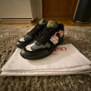Valentino Garavani Rockrunner sneakers - Säljer ett par Valentino Garavani Rockrunner sneakers med svart, rosa, grå och vit camouflage-mönster i storlek 42/43. Skorna har snörning, platt sula och rund tå. Materialmix av skinn och textil. Snygg detalj med logga på plösen och coola färgblock, kommer även med dustbag. Jag har endast använt det ett fåtal gånger förra året så det är bara några få småmärken av användning på de. Har du minsta fundering så är det bara att skriva🙌.‼️PRIS GÅR ATT DISKUTERA‼️