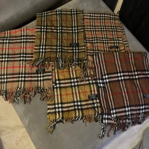 Rutig Burberry halsduk  - Säljer en klassisk rutig halsduk från Burberry i 100% lammull. Halsduk  har det ikoniska rutmönstret i beige, brunt och svart med inslag av rött. Fransen längs nederkanten ger en cool touch. Perfekt för dig som gillar tidlös design och vill sticka ut med ett lyxigt märke.