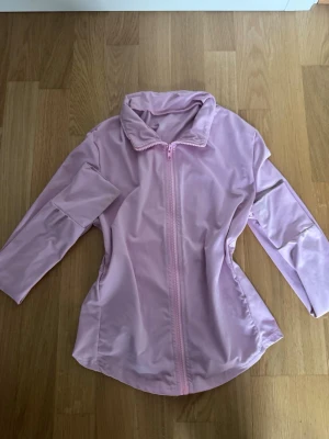 Ljusrosa vindjacka med dragkedja - Säljer en ljusrosa vindjacka med hel dragkedja framtill och hög krage. Jackan har en enkel, clean look och är tillverkad i ett lätt syntetmaterial. Perfekt för dig som vill ha en stilren jacka med sportig vibe.