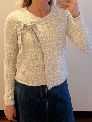 Odd Molly kofta med rosett - Superfin ljusgrå och vit kofta från Odd Molly med diskret mönster och omlott-design. Koftan har en snygg rosett vid axeln, två fickor fram och är tillverkad i 100% bomull. Perfekt för dig som gillar detaljer och mjuka material. Storlek 0/xs