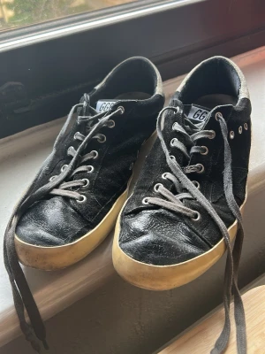 Golden Goose Superstar svarta sneakers - Svarta Golden Goose Superstar sneakers med beige glittrig häl och GGDB-detaljer. Skorna har snörning, rund tå och platt sula. Ovandelen är i mocka och skinn, med slitna kanter för en cool vintagekänsla. Sulan är beige och ger en snygg kontrast mot det svarta.