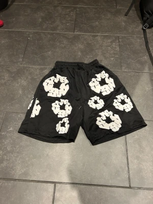 Svarta shorts från Denim Tears med tryck - Säljer ett par svarta shorts från Denim Tears med unikt vitt mönster som liknar kransar över hela plagget. Shortsen har elastisk midja med snörning och är gjorda i ett mjukt syntetmaterial som andas. Perfekt för dig som vill sticka ut med cool streetwear.