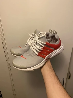 Nike Presto sneakers i grått och rött - Nike Presto sneakers med grå mesh-ovandel, vita snören och röd plastdetalj på sidorna. Snyggt röd swoosh på tån och AIR-tryck på hälen. Skorna har rund tå och platt sula, perfekt för dig som gillar sportig stil.