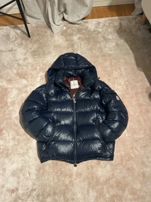 Mörkblå Moncler pufferjacka strl 1 - Säljer en mörkblå pufferjacka från Moncler med glansig finish och huva. Jackan har dragkedja framtill, två sidofickor med dragkedja och ett brunt innerfoder med Moncler-etikett och serietidningsdetalj. Perfekt för kalla dagar och riktigt snygg stil.