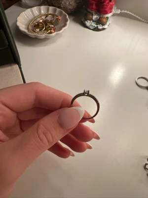 Snygg silverring med sten - En stilren silverfärgad ring med en gnistrande klar sten i mitten och små stenar längs bandet. Perfekt för dig som gillar klassiska smycken med lite extra bling. Ringen har en tunn och elegant form som passar till många olika stilar.