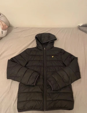  Lyle & Scott jacka - Svart pufferjacka från Lyle & Scott med huva och gul logotyp på bröstet. Jackan har quiltad design, dragkedja framtill och långa ärmar. Perfekt för kyliga dagar och har en clean, modern look.