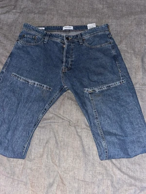 Blå jeans från Jack & Jones - Säljer ett par blå jeans från Jack & Jones i storlek W33. Jeansen har raka ben, klassiska fem fickor och unika horisontella sömdetaljer över låren. Tillverkade i slitstarkt denimtyg med normal passform och midja.