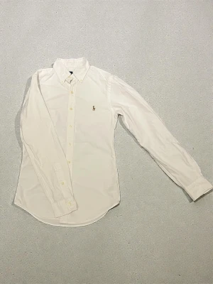 Vit slim fit skjorta från Ralph Lauren - Snygg vit skjorta från Ralph Lauren i slim fit-modell. Klassisk design med knappar framtill, lång ärm och broderad logga på bröstet. Tillverkad i mjuk bomull som känns skön mot huden. Perfekt för dig som gillar stilrena och tidlösa plagg.