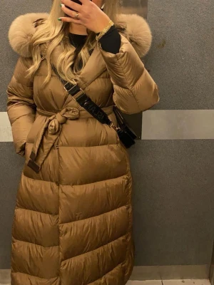 Brun lång pufferjacka med päls - Säljer en brun lång pufferjacka med fluffig päls runt huvan och bälte i midjan. Jackan har quiltad design och långa ärmar, perfekt för kalla dagar. Stängs med knappar och bältet runt midjan, saknar en knapp men finns extra knappar att sätta på från insidan av jackan. Köpt för 3899kr men säljer billigt pga knappen. Passform normal