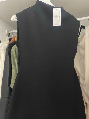 Svart ärmlös klänning med hög krage - Snygg svart klänning från primark med stilren, rak passform och hög krage. Klänningen är ärmlös och har en minimalistisk design som gör den enkel att matcha med olika accessoarer. Perfekt för dig som gillar en clean och modern look.